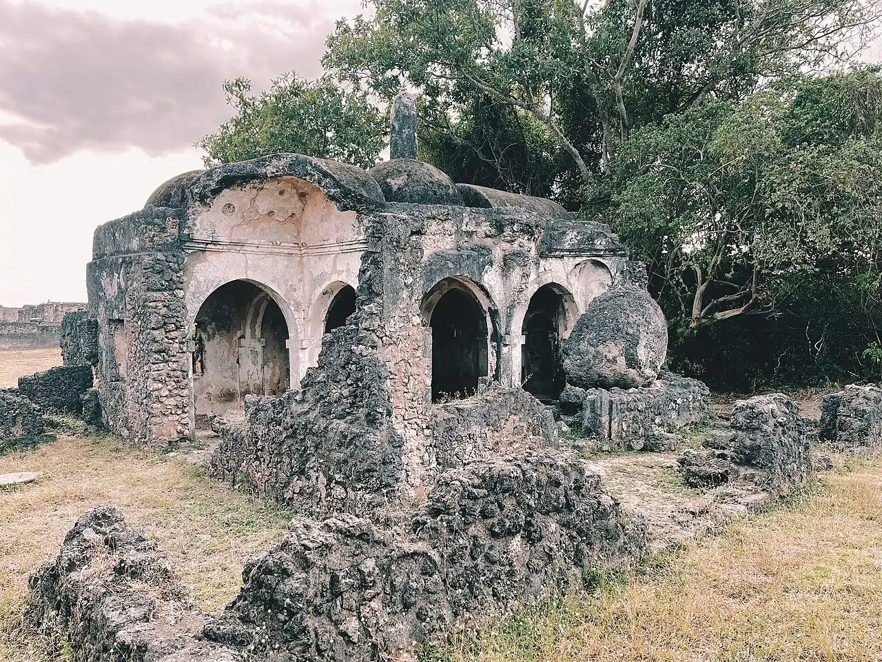The surviving arches and domed structure of a small mosque among vegetation at Kilwa Kisiwani.