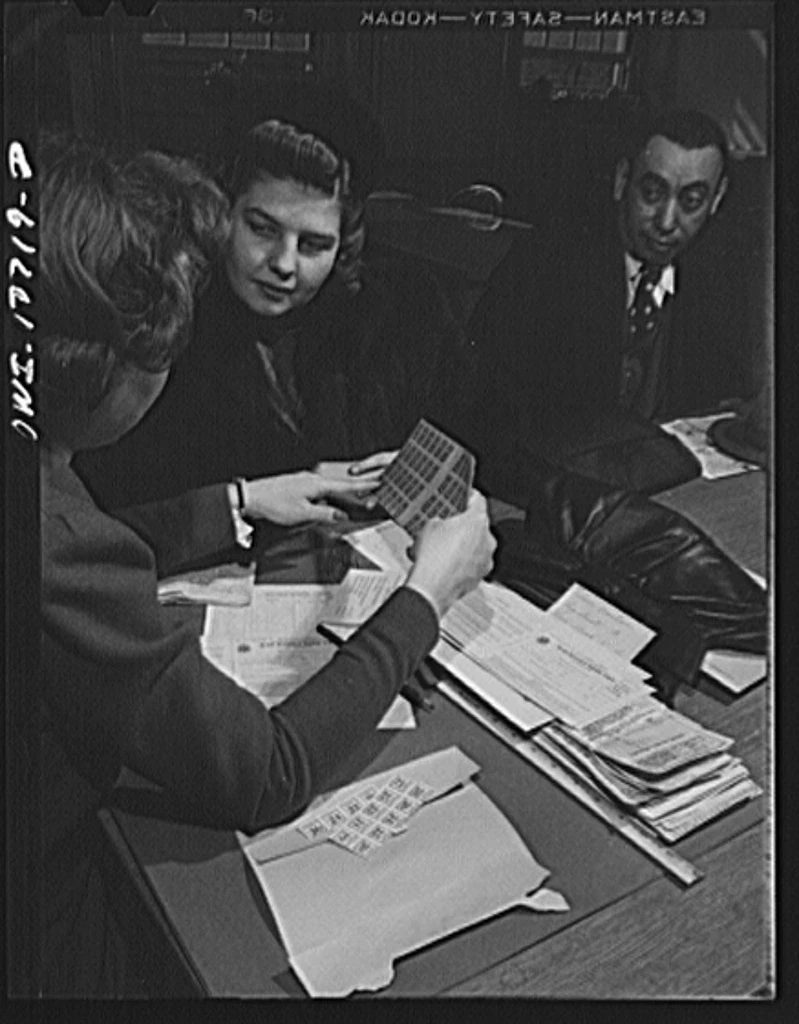A woman holds up ration coupons at a registration desk while two people listen.