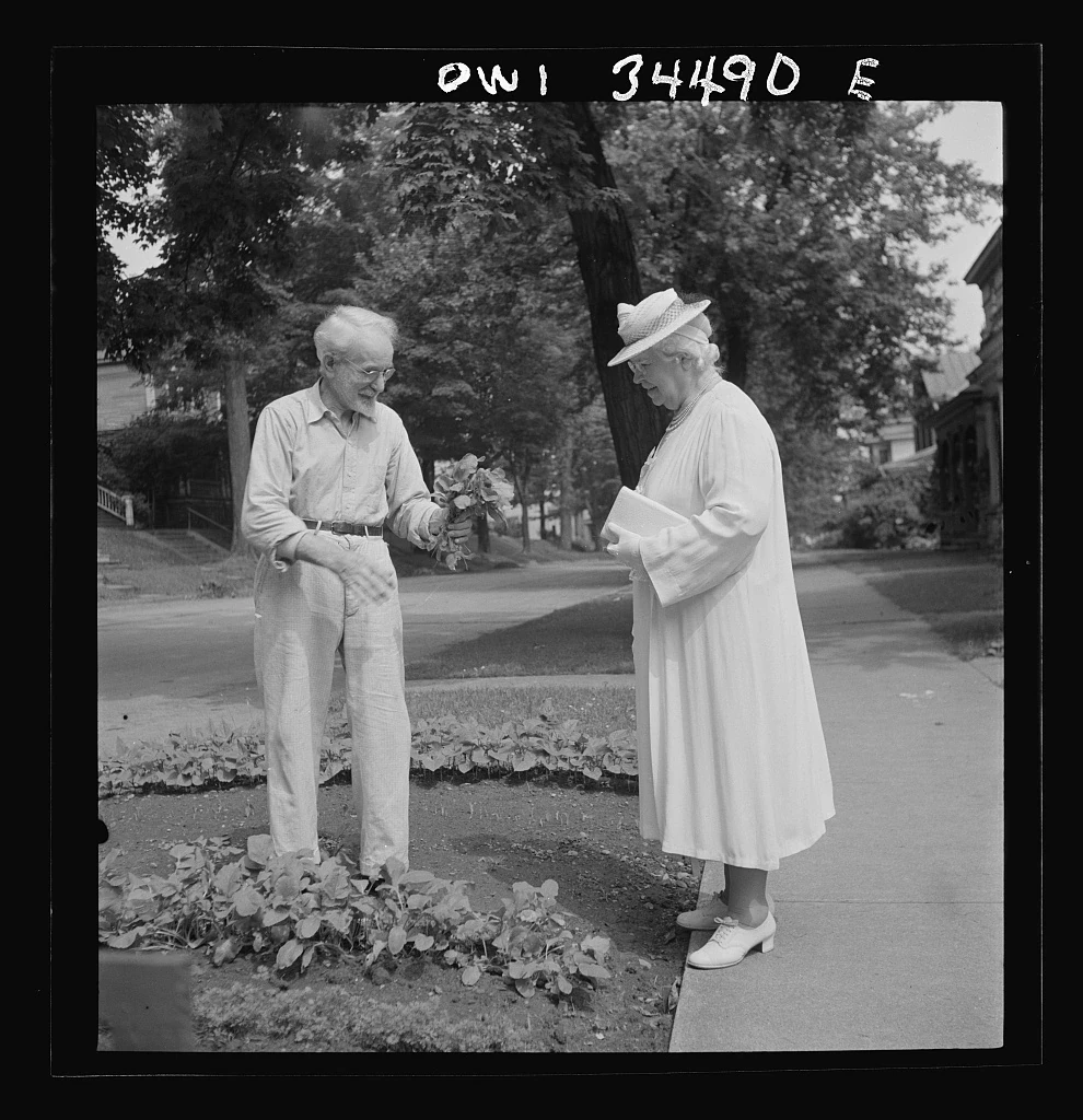 A man holds freshly picked vegetables beside a woman standing near a small front garden.